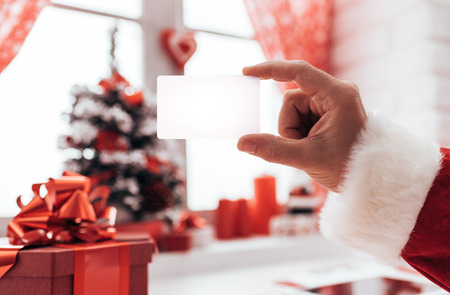 Santa Claus holding a blank business card, window and Christmas decorations on the backgroundの写真素材