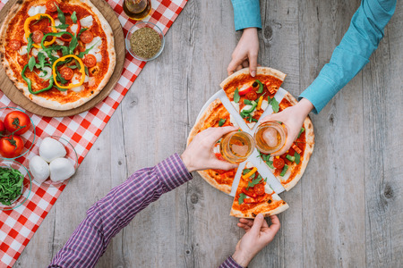 Friends eating a tasty pizza together at the restaurant, they are holding a slice and toasting with beerの写真素材