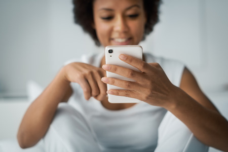 Young smiling woman sitting on the bed at home and texting with her smartphone, she is social networking and chattingの写真素材