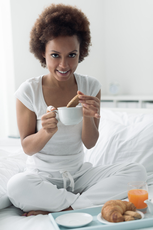 Young african american woman having a relaxing breakfast in bed, she is dipping cookies into a cup of teaの写真素材