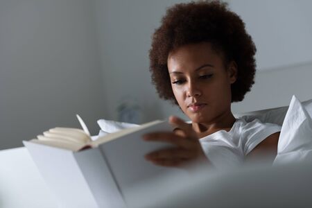 Young african woman relaxing at home and reading a book, she is lying on the bedの写真素材