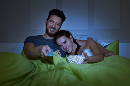 Happy couple watching films on tv in bed, the man is holding a remote control and the woman is eating popcornの写真素材