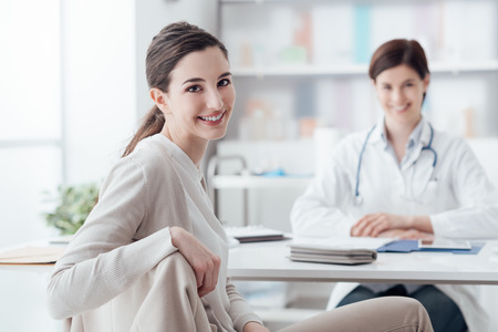 Smiling patient receiving a medical consultation and looking at camera, the female doctor is sitting at desk on the backgroundの写真素材