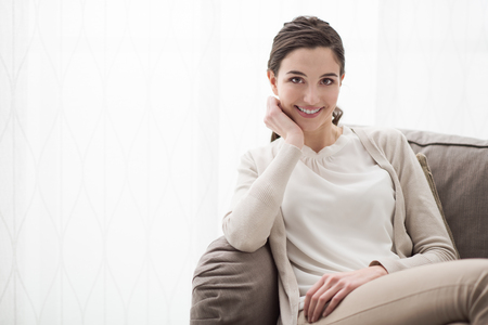 Smiling young girl posing on the couch, she is relaxing and looking at camera with hand on chinの写真素材