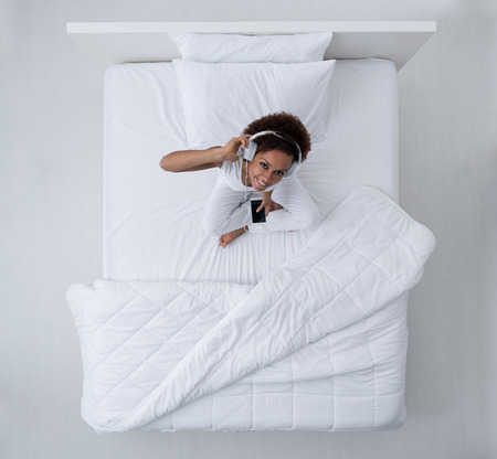 Cheerful young mixed race woman relaxing in bed, she is listening to music using a smartphone and headphones, top viewの写真素材