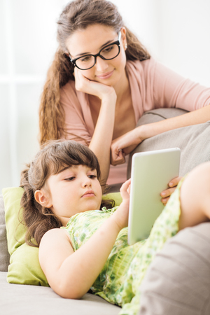 Smiling young mother and daughter using a digital tablet together at home, the girl is using apps for kidsの写真素材