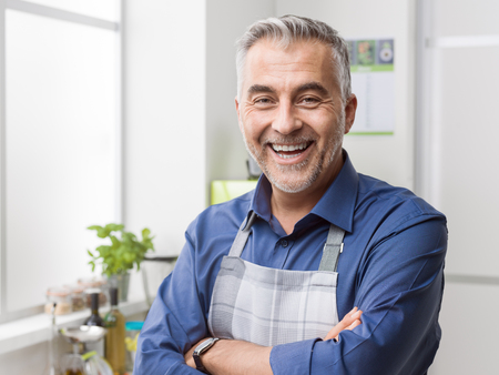 Confident smiling man posing in his kitchen, he is wearing an apron and posing with arms crossedの写真素材