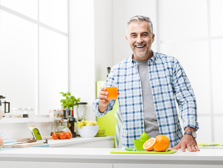 Smiling man having breakfast at home, he is preparing and drinking a glass of fresh orange juice, kitchen interior on the backgroundの写真素材