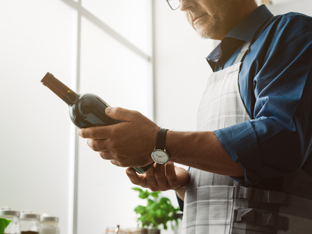 Man holding a red wine bottle and reading the label, he is cooking in the kitchen at homeの写真素材