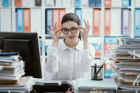 Confident secretary sitting at office desk and adjusting her glassesの写真素材
