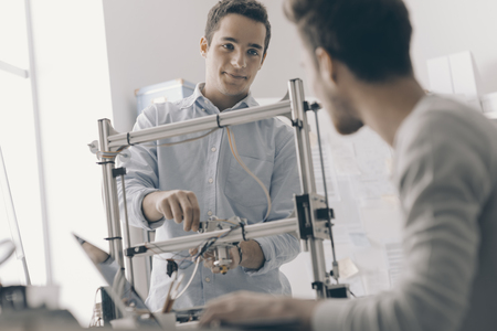 Engineering students working in the lab, a student is adjusting a 3D printer's components, the other one on foreground is using a laptopの写真素材