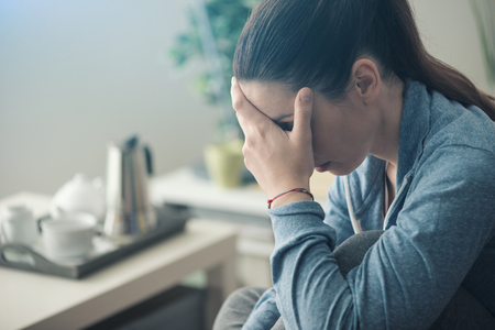 Sad depressed woman at home sitting on the couch, looking down and touching her forehead, loneliness and pain conceptの写真素材
