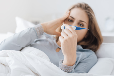 Young woman lying in bed with flu virus, she is measuring her temperature with a thermometer and touching her templeの写真素材