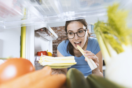 Young smiling woman preparing an healthy fresh meal and eating celery, fridge interior and groceries on the shelvesの写真素材