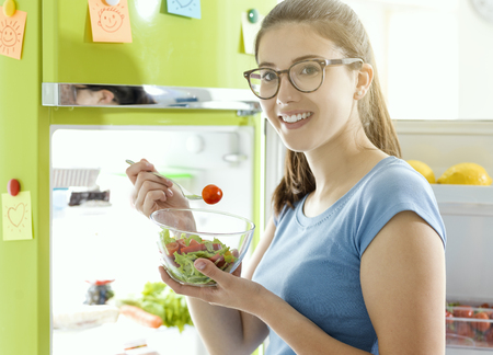Smiling woman eating some fresh salad next to the fridge, healthy vegan food and dieting conceptの写真素材