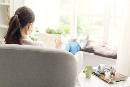 Young woman relaxing at home next to a window, she is sitting on the armchair and reading a bookの写真素材