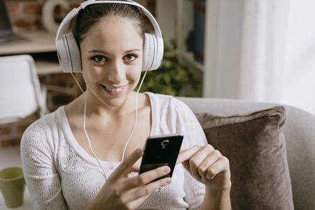 Happy young woman listening to music at home, she is connecting with her smartphone and wearing headphonesの写真素材