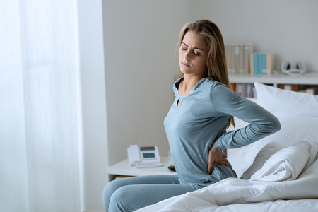 Young woman with back ache, she is sitting on the bed and touching her backの写真素材