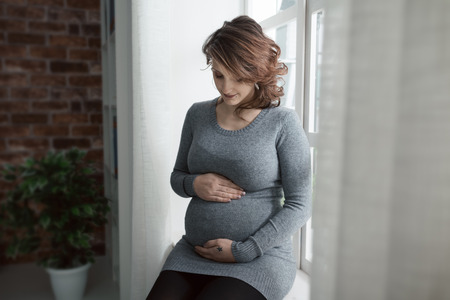 Young pregnant woman relaxing at home and smiling, she is sitting on the windowsill, motherhood conceptの写真素材