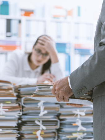 Exhausted young secretary overloaded with work, her boss is bringing more paperwork to her