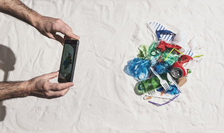 Man taking a picture of waste pollution on the beach and sharing it online using his smartphone,...