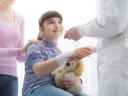 Doctor wrapping a girl's injured wrist with gauze and bandage, the mother is standing next to the daughter, first aid and healthcare conceptの写真素材