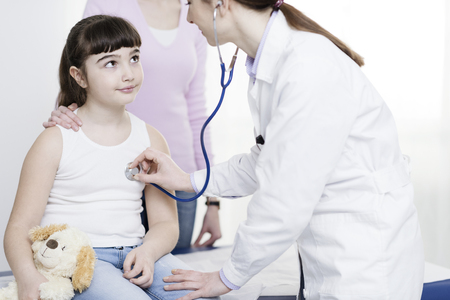 Female doctor examining a cute smiling girl with a stethoscope, the mother is next to her, children and healthcare conceptの写真素材