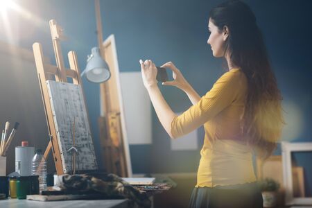 Young woman in the art studio taking a picture of her painting using her smartphone, art and...