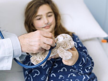 Friendly doctor assisting a kid at the hospital, he is checking her plushie's heartbeat