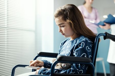 Cute girl in wheelchair at the hospital using a tablet, her mother and the doctor are talkingの写真素材