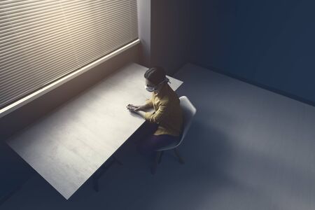 Quarantined lonely woman with covid-19 wearing a face mask and sitting at home alone in front of a window, virus outbreak emergency conceptの写真素材