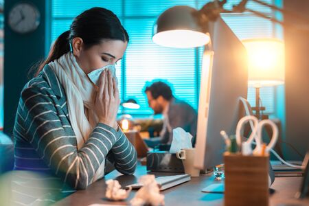 Young office worker sitting at office desk and working, she is having a cold and blowing her nose with a tissueの写真素材