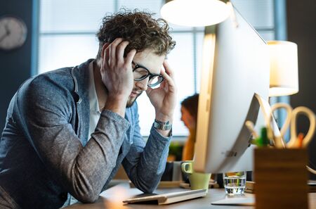 Stressed office worker sitting at desk and working with his computer, he is tired and having a headacheの写真素材
