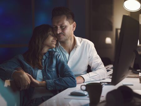 Happy loving couple relaxing at home late at night and hugging, they are sitting at desktop togetherの写真素材