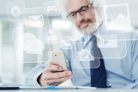 Smiling businessman sitting at office desk and using a touch screen smart phone, texting and...