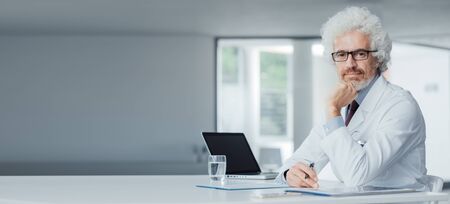 Confident doctor with hand on chin sitting at office desk and posing, hospital interior on backgroundの写真素材