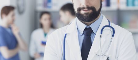 Confident young doctor posing in the office, he is smiling at camera, medical team on the backgroundの写真素材