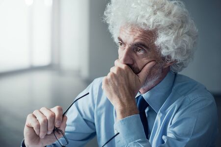 Pensive confused businessman with hand on chin sitting at office desk and looking awayの写真素材