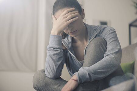 Sad depressed woman at home sitting on the couch, looking down and touching her forehead, loneliness and pain conceptの写真素材
