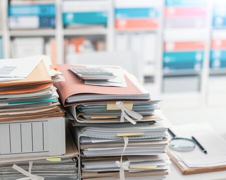 Stacks of files and paperwork in the office and bookshelves on the background: management and...