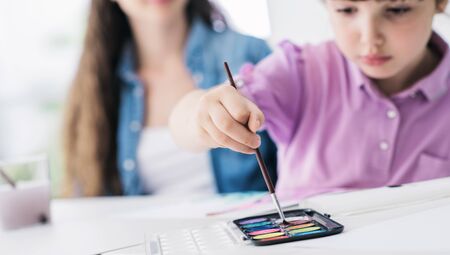 Mother and child spending time together at home, the girl is painting with brush and watercolors