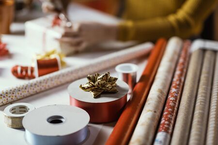 Woman wrapping Christmas gifts at home, she is tying a ribbon bow: holidays and celebrations concept