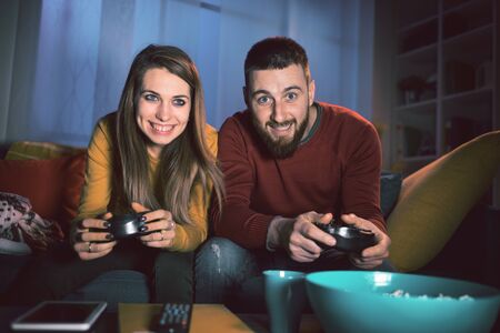 Happy couple playing video games together at home, they are sitting on the sofa and holding a game controller, entertainment and lifestyle conceptの写真素材
