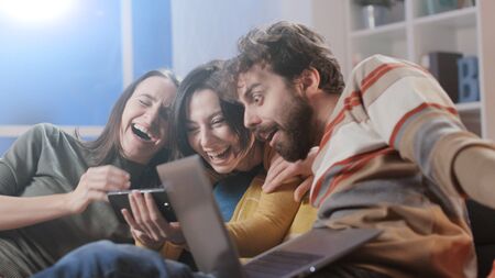 Happy friends sitting on the sofa at home and social networking with their smartphone, togetherness and technology conceptの写真素材