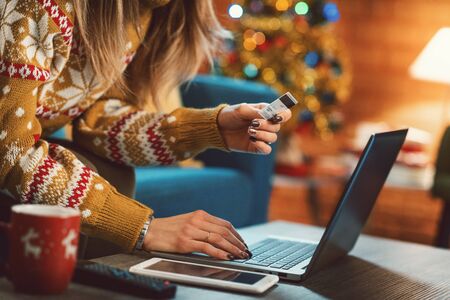 Woman doing online shopping at Christmas: she is sitting on the sofa at home, connecting with her laptop and holding a credit card, Christmas tree in the backgroundの写真素材