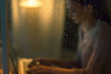 Young woman working with a laptop next to a window on a rainy night, glass with raindrops in the...