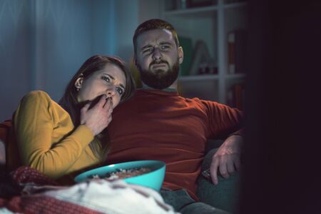 Young couple sitting on the couch at home and watching horror movies together on TV, he is hugging his girlfriendの写真素材
