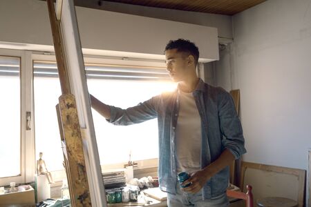 Young creative artist working on his artwork in the studio, he is standing and painting on a canvas