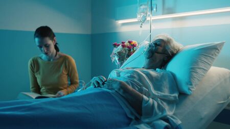 Woman at the hospital assisting a senior patient with oxygen mask at night, she is reading a book and checking herの写真素材