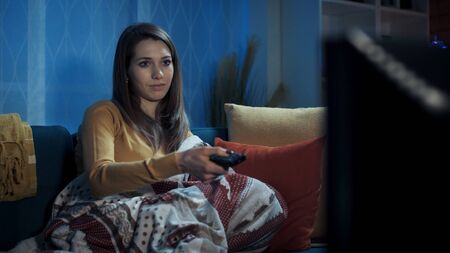 Serene woman sitting on the sofa at home and watching television, she is holding the remote control...
